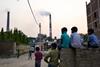 An image showing boys sitting and watching street cricket as emissions billow from smokestacks at the NTPC Ltd. Badarpur coal-fired power plant near residential property in Badarpur, Delhi, India