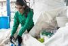 A black-haired woman in a green long-sleeve shirt, black rubber gloves and lab specs is picking plastic bottles out of a huge bag of empty bottles. There are more such bags piled next to her.