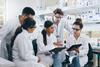 A group of young male and female scientific researchers in a lab and wearing lab coats and safety glasses gather around to discuss something on a computer tablet.