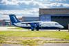The FAAM research aircraft outside its hangar at Cranfield airport. It is a blue and white research aircraft parked on the apron in front of its hangar. There is green grass in front of the aircraft and blue sky with clouds in the background.