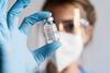 A photograph of a researcher wearing eye-protection, gloves and a mask, holding a vial containing an mRNA vaccine