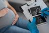 Pregnant woman in the background sitting on a couch while a doctor looks at an ultrasound image of a fetus on a clipboard in the foreground.