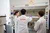 A member of staff at a laboratory instructs some students on how to use a reaction set-up in a fume cupboard