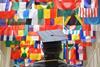 A man in a mortar board and graduation gown looks up at flags from countries all over the world.