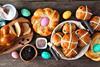 Hot cross buns in a basket sit on a table amongst other well-known, delicious Easter breads, along with some ingredients including raisins