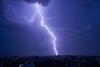 Powerful bolt of lightning cutting through dark, stormy sky over cityscape at night. Lightning's bright illuminate scene, contrasting sharply with dark clouds and dim lights of buildings below