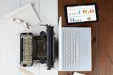 Image shows a laptop and old fashioned typewriter plus various desk items