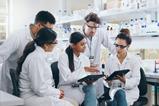 A group of young male and female scientific researchers in a lab and wearing lab coats and safety glasses gather around to discuss something on a computer tablet.