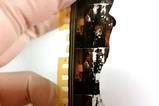 Close up of a damaged strip of celluloid film held by a hand in latex gloves. The film features two women in mid-century living room and clothing but the film is cracked and broken and parts of the images have turned white.