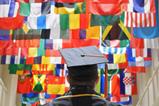 A man in a mortar board and graduation gown looks up at flags from countries all over the world.