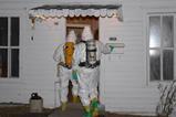 Two figures in white hazmat suits with breathing apparatus enter through the door of a rundown house