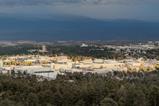 An aerial view of Los Alamos National Laboratory in the USA. There a number of low-rise office-type buildings surrounded by forest with cloud-topped mountains in the distance