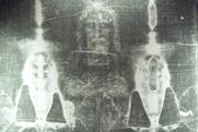 An enhanced black and white image of the Turin Shroud featuring a faint image of a bearded man's face some a damaged cloth