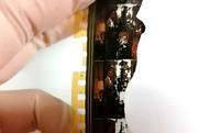 Close up of a damaged strip of celluloid film held by a hand in latex gloves. The film features two women in mid-century living room and clothing but the film is cracked and broken and parts of the images have turned white.
