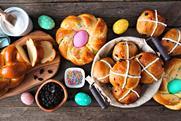 Hot cross buns in a basket sit on a table amongst other well-known, delicious Easter breads, along with some ingredients including raisins