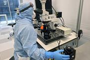 A scientist in full PPE looks through the scope on a microscope