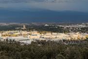 An aerial view of Los Alamos National Laboratory in the USA. There a number of low-rise office-type buildings surrounded by forest with cloud-topped mountains in the distance