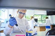 A female scientist using a pipette at a laboratory workbench. She is wearing a lab coat, safety goggles and latex gloves. Behind her other scientists are working at a centrifuge