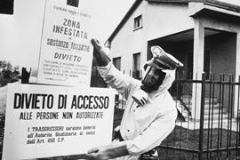 A black and white image of a policeman in Seveso, Italy pinning up warning signs written in Italian warning of chemical contamination of the area