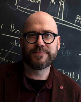 Portrait of Alan Aspuru-Guzik, principle investigator at El Agente, pictured in front of a blackboard