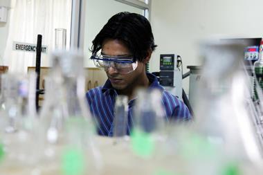 A photo showing a dark-haired man wearing lab safety goggles over a pair of regular glasses. He's wearing a blue striped shirt and tie and focuses a concentrated look at something on the bench in front of him.