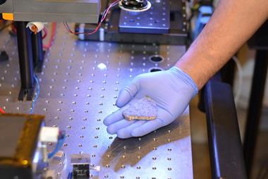 A photo of a flat rock fragment held in the palm of a purple-gloved hand. The person's hand rests on what looks like a work bench surrounded by scientific electronic equipment