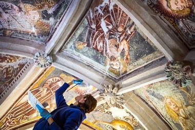 A photo shows a woman wearing a blue lab coat and blue nitrile gloves concentrating on using a brush to touch up a large fading ceiling fresco