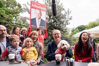 A white, middle-aged, bespectacled man seated at a table. He's laughing and holding a dog on his lap. There is a small crowd of people gathered to his right, one holding an election campaign poster.