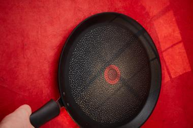 A photo shot from above of someone's hand holding the handle of a non-stick frying pan. The pan is sitting on what looks like a table with a red tablecloth.