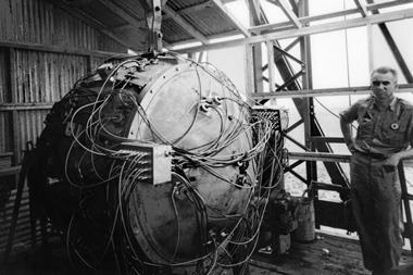 A black-and-white photo showing Norris Bradbury next to the Trinity test atom bomb. The device consists of a metal casing, a bit taller than Bradbury, with cables coming out of various places.