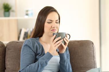 A white woman with brown hair wearing a white shirt and blue cardigan, sitting on a brown sofa chair. She's smelling a cup of coffee, a deeply disgusted look on her facecoffee