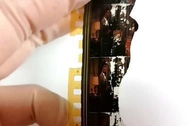 Close up of a damaged strip of celluloid film held by a hand in latex gloves. The film features two women in mid-century living room and clothing but the film is cracked and broken and parts of the images have turned white.