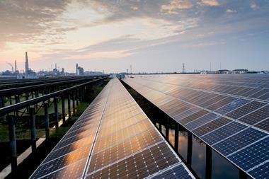 A photo of solar panels during sunset on a bright day. There's an industrial complex in the far distance
