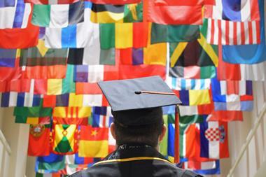 A man in a mortar board and graduation gown looks up at flags from countries all over the world.