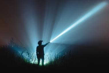 A photo of a person's silhouette standing with the back to the viewer. They're holding a torch that casts a bright blue beam of light across the dark landscape.