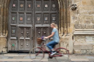 A photo of an large wooden gate featuring several coats of arms. It's the entrance door to one of the colleges at the University of Oxford. A person on a bike is cycling past, the motion blurring the details