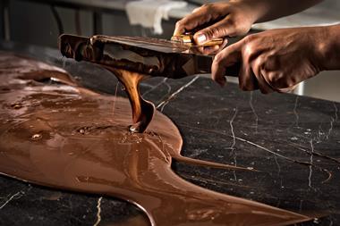 A photo of a person's hands holding two long spatulas covered in melted chocolate. The chocolate is dripping off the spatulas and pooling on a marble surface underneath, which is covered in more molten chocolate.
