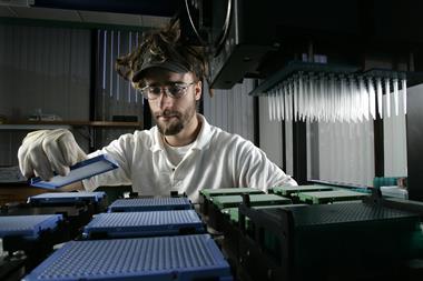person in a white shirt with a beard and glasses places a blue pipette rack next to some others. To their right a grid of pipettes dangles down from a liquid dispensing robot