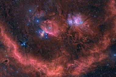 An image showing the Orion stellar neighbourhood as red-glowing dust clouds and hundreds of tiny dots of newly born stars