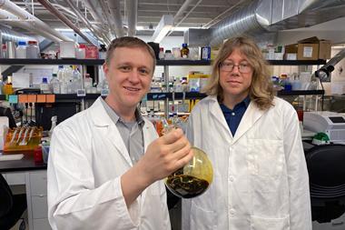 Two scientists in lab coats in a modern lab. One is holding a glass flask with a dark goo inside.
