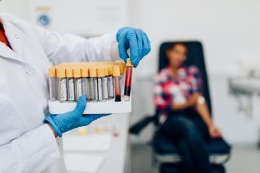 A medical professional with a tray of blood samples and a test subject behind holding a dressing to her arm where she has had a blood sample taken.