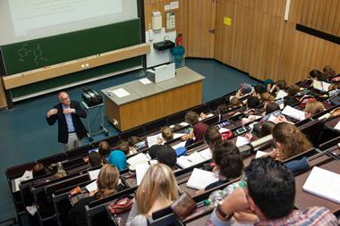 A man stands at the front of a lecture theatre in front of several rows of students sat with papers on the benches in front of them. On the blackboard behind him is a chemical structure