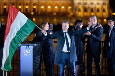 Peter Magyar standing on a stage waving a Hungarian flag while his supporters embrace each other behind him. On the other side of the River Danube you can see the Hungarian parliamentary building lit up at night.