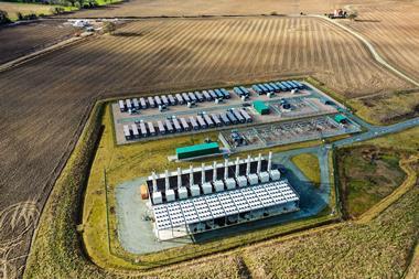 An aerial view of a clean industrial site of dark storage containers and a building with lots of chimneys. It is surrounded by freshly ploughed farmland.