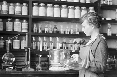 A black-and-white photo showing an early 20th century lab. A woman wearing an apron is concentrating on retrieving something out of a desiccator with a pair of tongs, holding the lid with her other hand. Behind her, shelves are lined with chemical bottles