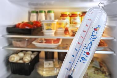 A close-up photo of a plastic fridge thermometer that shows a temperature close to 0C. In the background, an open fridge shows shelves filled with food.