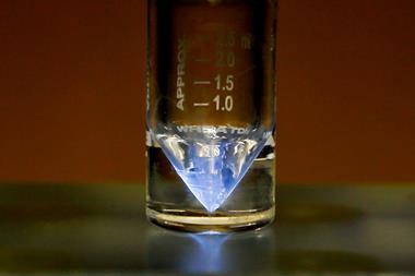 A photo showing a closeup of a small glass vial with a black plastic screw-on top. At the bottom of the pointed vial sits a small amount of blueish liquid that has a glowing shine to it.