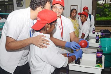 A scientist helps two visually impaired high school students perform a chemistry experiment with a bottle of fizzy drink and a balloon. The students are smiling as they feel the changes to the objects.