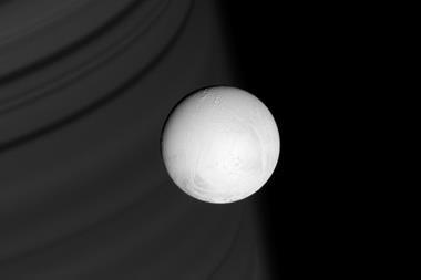 An image of Enceladus, a cratered white sphere hanging in the dark of space. Saturn's rings can be seen as faint grey stipes across the background of the photo