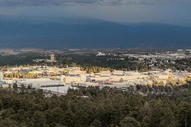An aerial view of Los Alamos National Laboratory in the USA. There a number of low-rise office-type buildings surrounded by forest with cloud-topped mountains in the distance